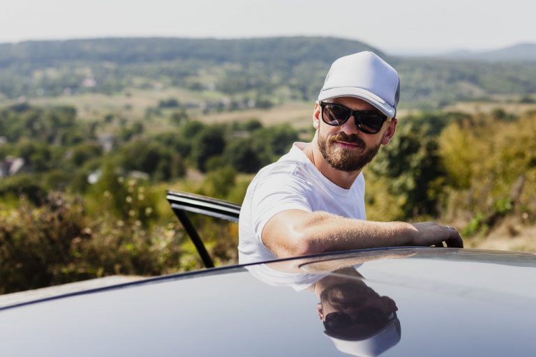 Man wearing baseball cap and sunglasses leaning against the roof of a car.