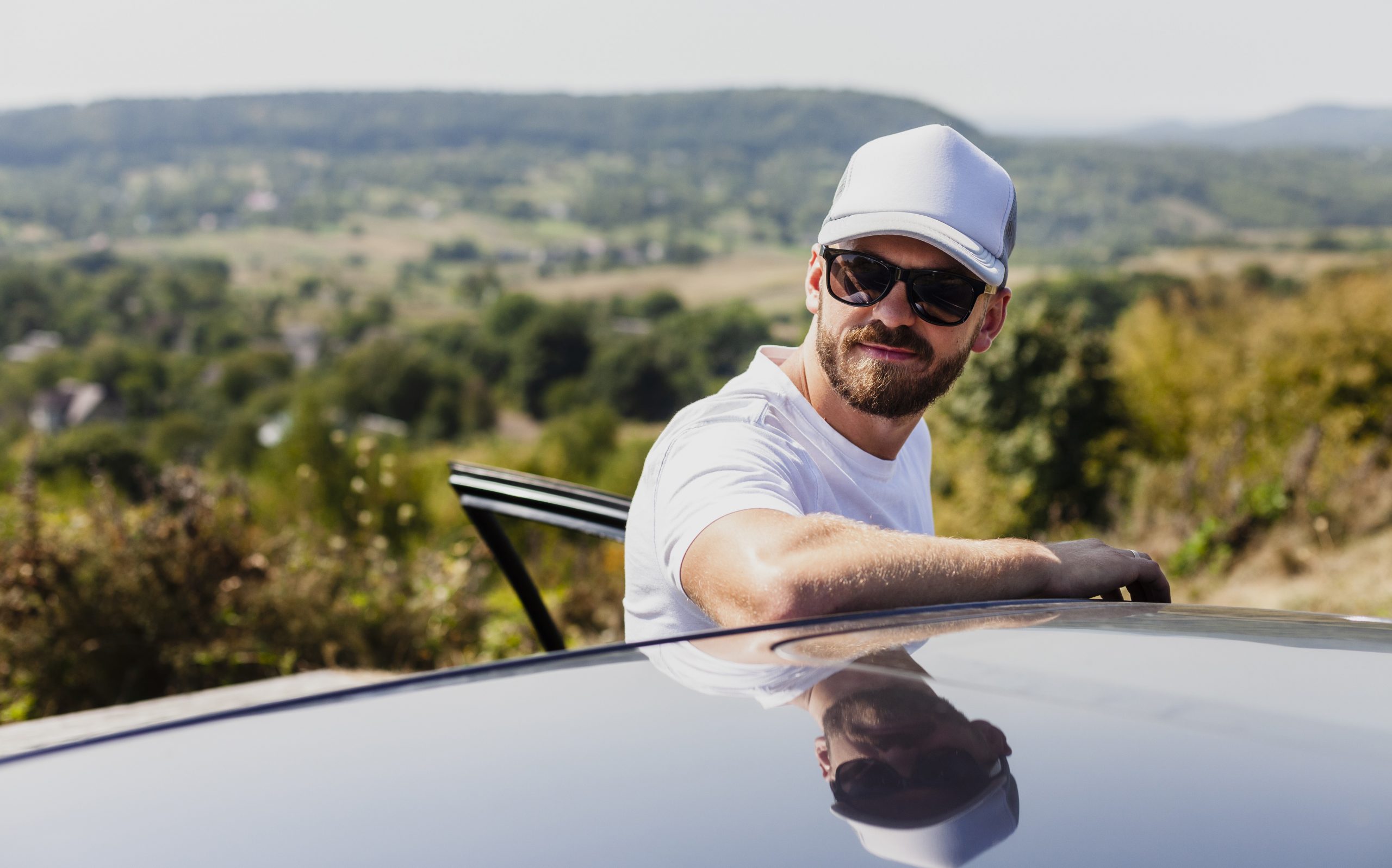 roof-car-fp Man wearing baseball cap and sunglasses leaning against the roof of a car.